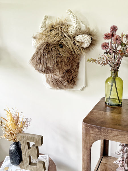 A plush Highland Cow Wall Mount handmade by ClaraLoo in a rustic western themed nursery. This stuffed highland cow is made of soft brown faux fur with crocheted ears and crocheted horns. This plush highland cow is mounted on a white wooden plaque.