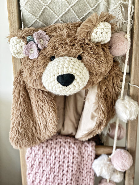 A plush brown bear rug made by ClaraLoo with crochet muzzle and ears, placed on a wooden ladder with a knit patterned blanket.