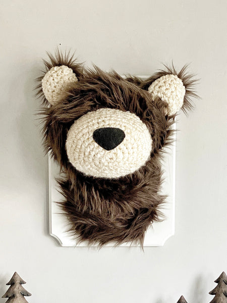 A plush bear head mounted on a white wooden plaque in a forest themed nursery. This stuffed bear wall mount is handmade by ClaraLoo with soft brown faux fur and crocheted accents.