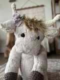 Stuffed highland cow toy with crochet horns, ears, and feet. The highland cow is made with tan fur and has purple crochet flowers attached to a horn.