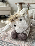 Plush highland cow in a kneeling position on a carpet. The highland cow toy is made with tan fur and has crochet horns, ears, and feet.
