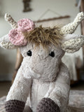 Stuffed highland cow with tan fur and crochet horns, ears, and feet. The highland cow has a pink crochet bow attached to the horn.