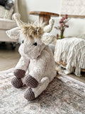 Stuffed highland cow sitting on a carpet in a bedroom. The plush highland cow has tan fur and crochet horns, ears, and feet.