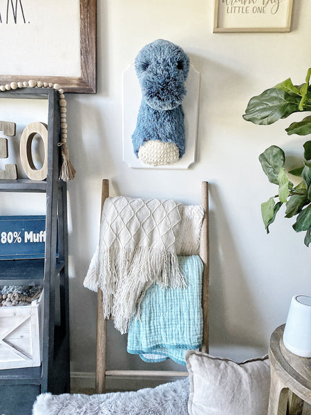 A plush Brontosaurus Wall Mount made by ClaraLoo in a dinosaur themed nursery. The stuffed dinosaur is made of soft blue faux fur with crocheted accents. The plush dinosaur is mounted on a white wooden plaque.