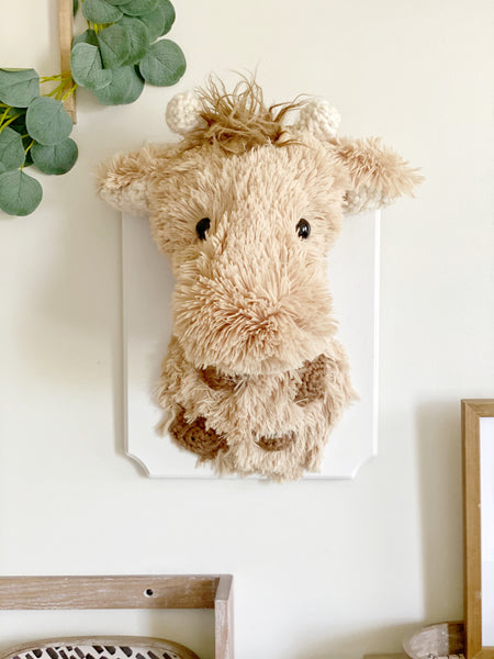 A plush giraffe wall mount handmade by ClaraLoo in a safari themed nursery. This stuffed giraffe is made of tan faux fur with crocheted horns, ears, and spots. The plush giraffe is mounted on a white wooden plaque.