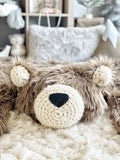A handmade ClaraLoo bear rug with brown faux fur and cream colored ears and face. The bear's ears and face are crocheted. The bear has a black nose and is laying on a shaggy cream colored carpet.