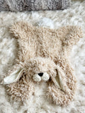 A tan bunny lovey made by ClaraLoo with a plush head, white tail, and crochet muzzle, lying on a fluffy white carpet.