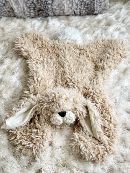 A tan bunny lovey made by ClaraLoo with a plush head, white tail, and crochet muzzle, lying on a fluffy white carpet.