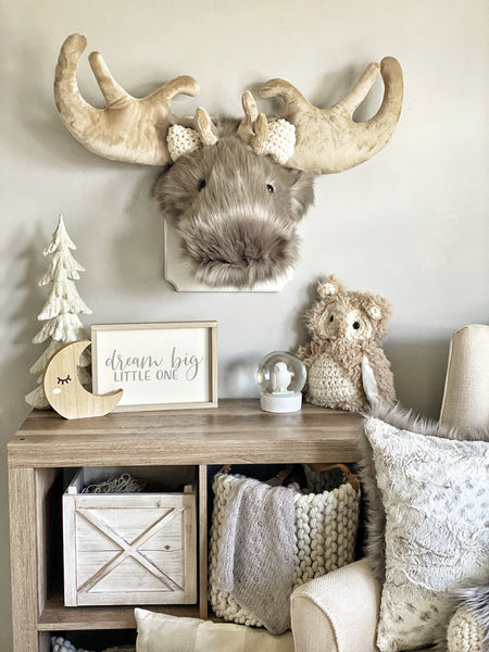 A stuffed moose head mounted on a white wooden plaque in a woodland themed bedroom. This plush moose wall mount is handmade by ClaraLoo with soft gray faux fur and crocheted ears.